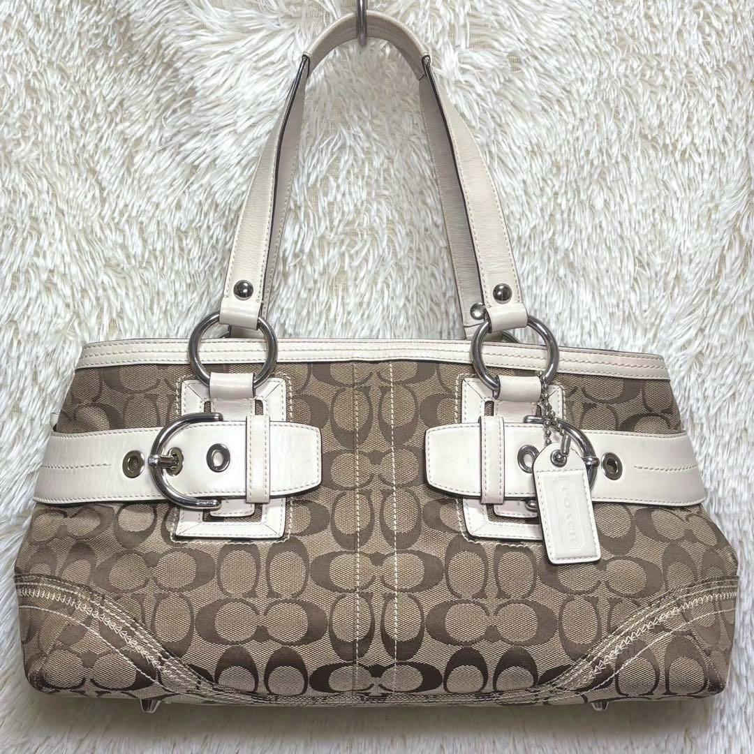 Front view of a COACH Soho Signature tote bag in brown signature canvas with white leather trim and a hanging COACH tag.