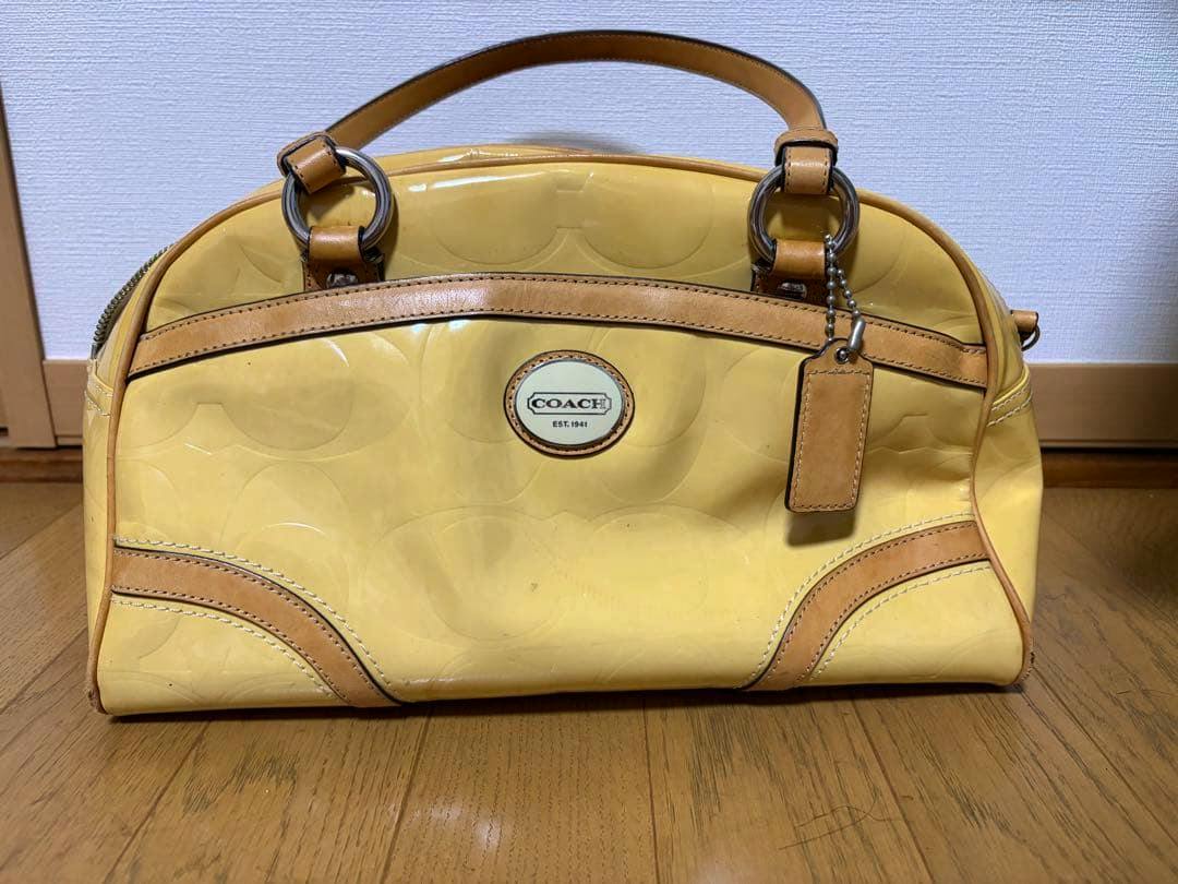 Front view of a yellow Coach patent leather handbag with leather trim, top handles, metal rings, and a hanging leather tag.