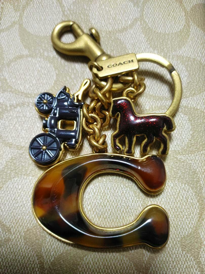 Close-up of a COACH bag charm showing a large tortoiseshell-style letter C, a horse and carriage charms, and a gold-tone clasp.