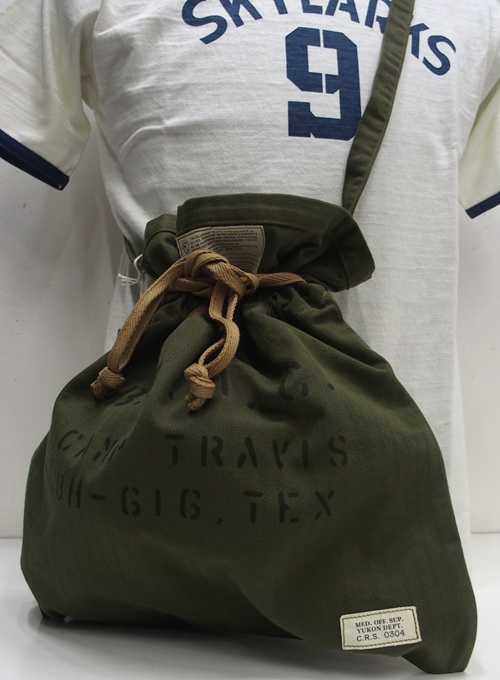 Olive drab COLIMBO Clara Bag (40's Apron Bag) worn crossbody, showing drawstring closure, stenciled text, and a sewn label.