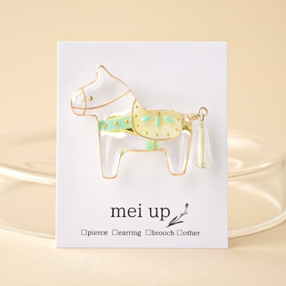 A clear horse-shaped brooch with gold metal outline and pale green saddle pinned to a white display card.
