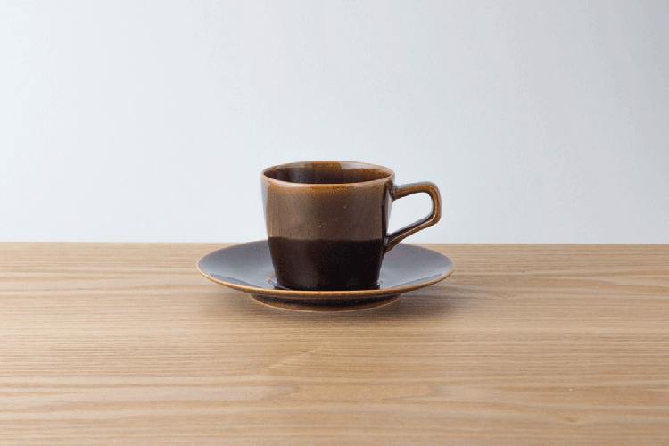 A brown glazed ceramic cup and matching saucer on a wooden surface against a white background.