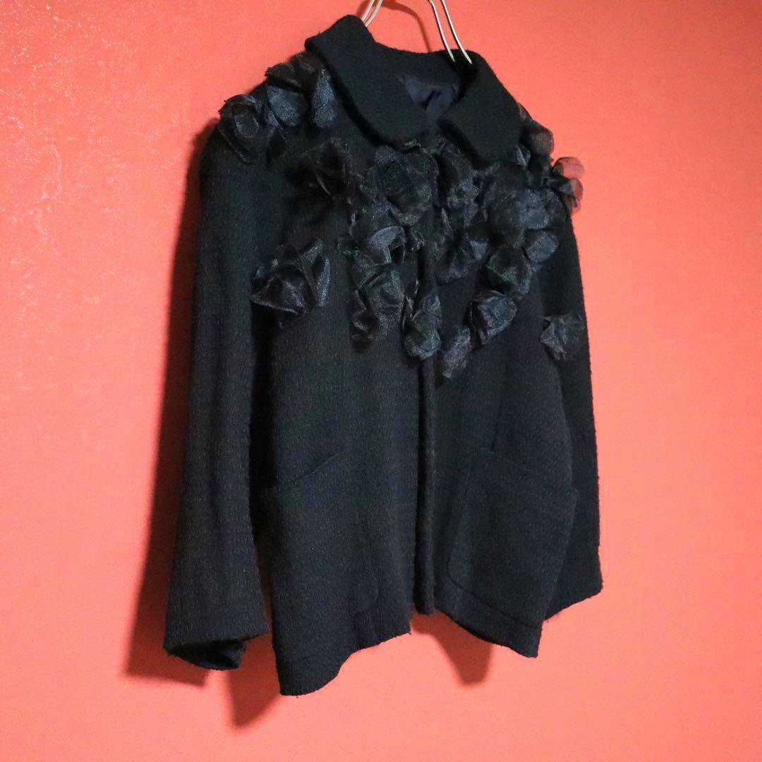 Black COMME des GARÇONS round-collar coat with decorative fabric details hanging on a hanger against a red background.