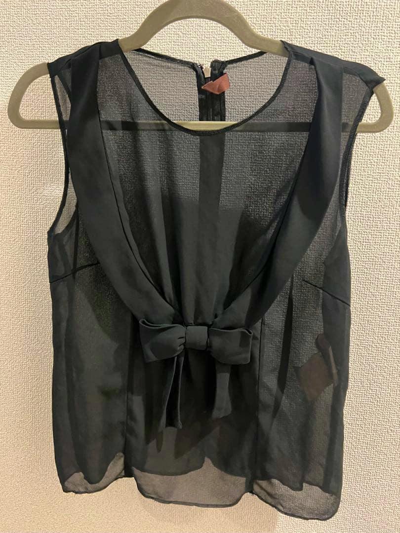 Black chiffon sleeveless blouse with a front ribbon bow shown hanging on a hanger against a textured wall.