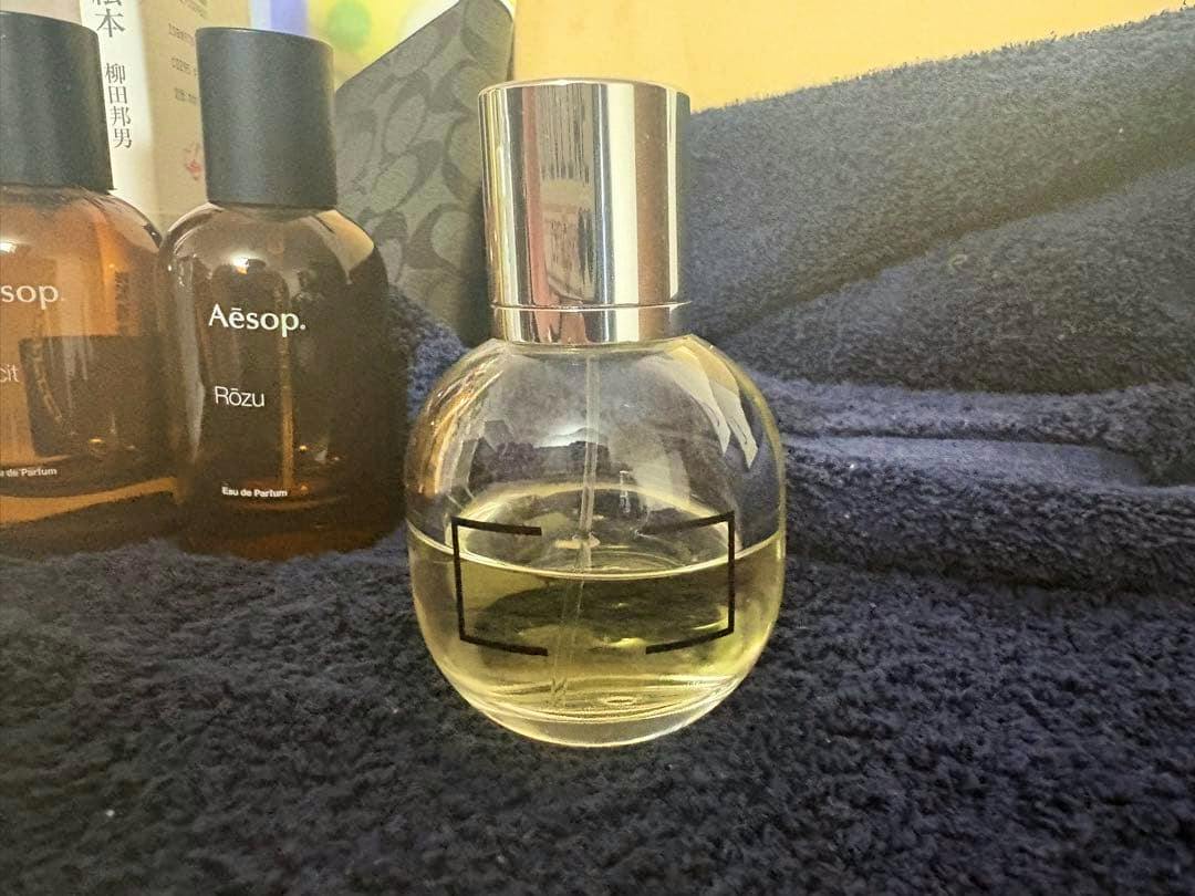 A round glass perfume bottle with a silver cap and yellow liquid sitting on a dark cloth with two brown bottles visible behind it.