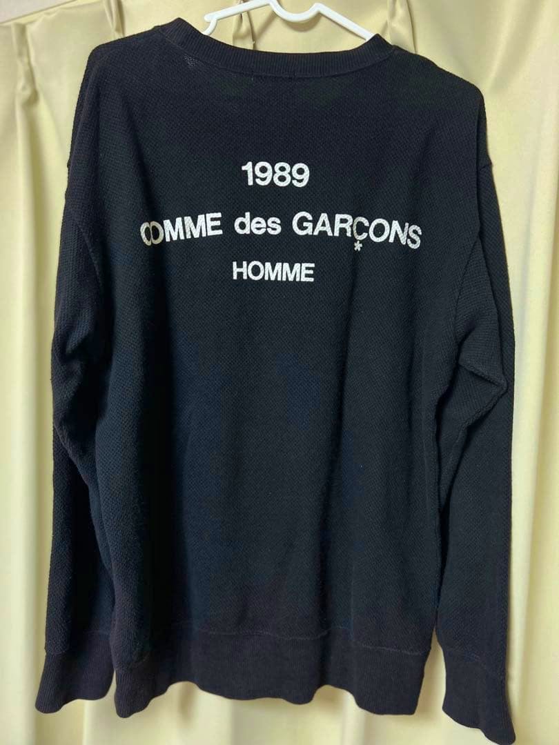 Black COMME des GARÇONS Homme cotton sweatshirt showing printed "1989 COMME des GARÇONS HOMME" on the back, hanging on a hanger.