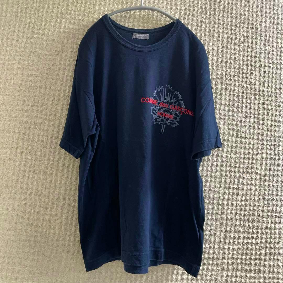 Front view of a navy COMME des GARCONS HOMME Tシャツ (00s) with a red logo and graphic, hanging on a hanger against a textured wall.