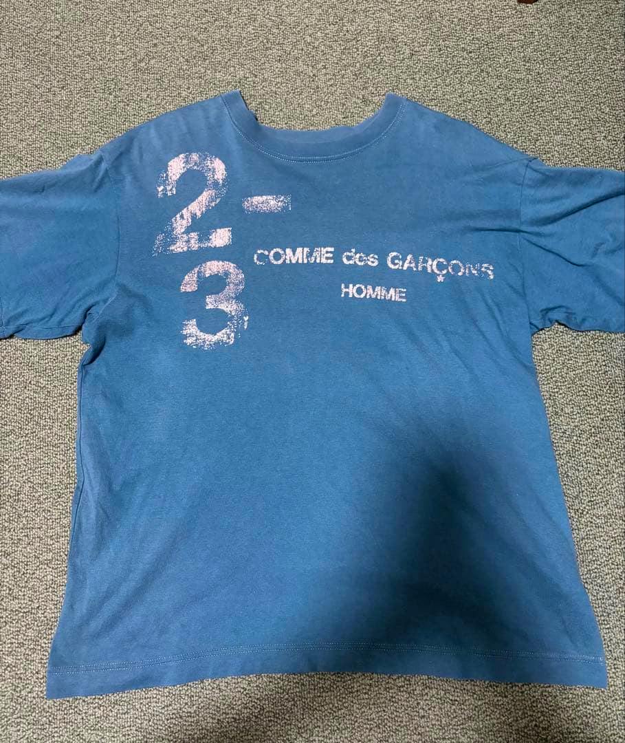Front of a blue men’s T-shirt laid flat showing a distressed "2-3" print and the "COMME des GARÇONS HOMME" logo on the chest.