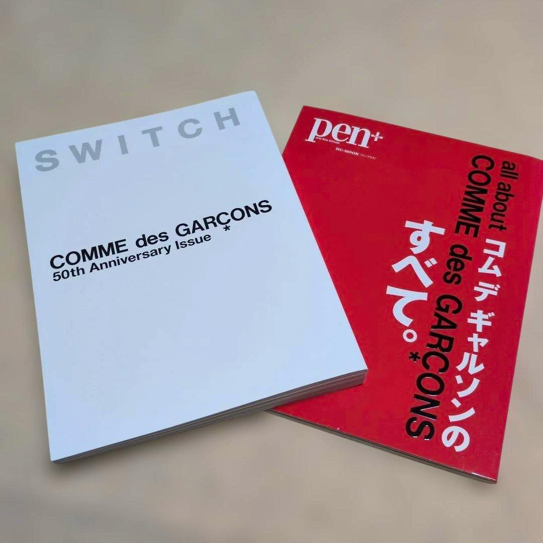A white magazine titled "COMME des GARÇONS 50th Anniversary Issue" lies next to a red magazine showing vertical "COMME des GARÇONS" text.