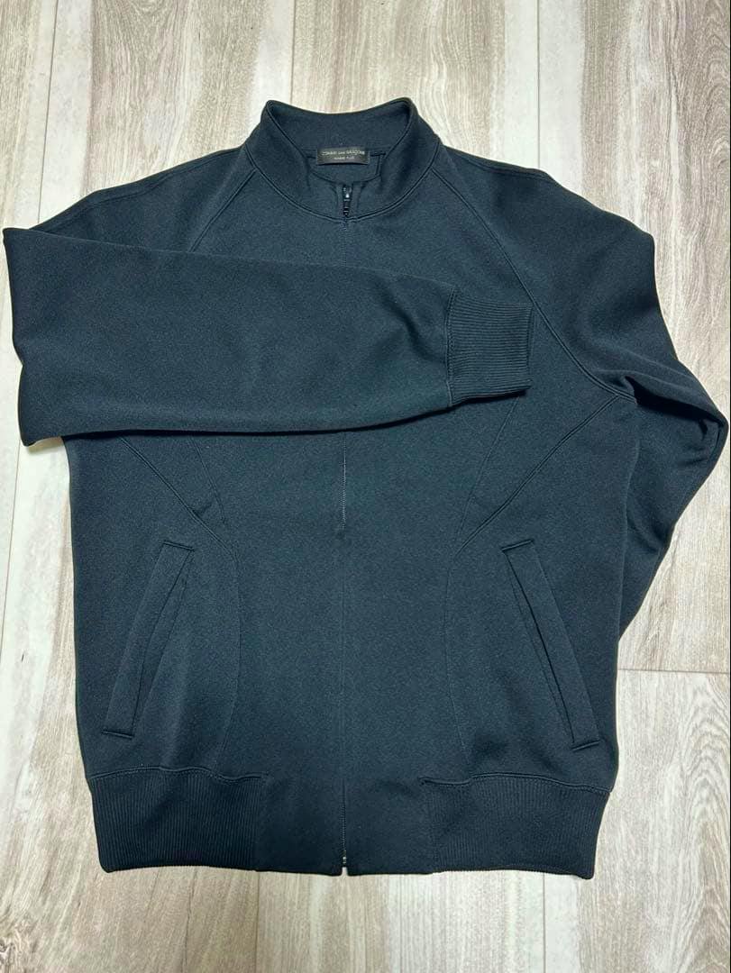 Black men's track jacket laid flat on a wooden floor showing the front zipper, pockets, and ribbed hem.