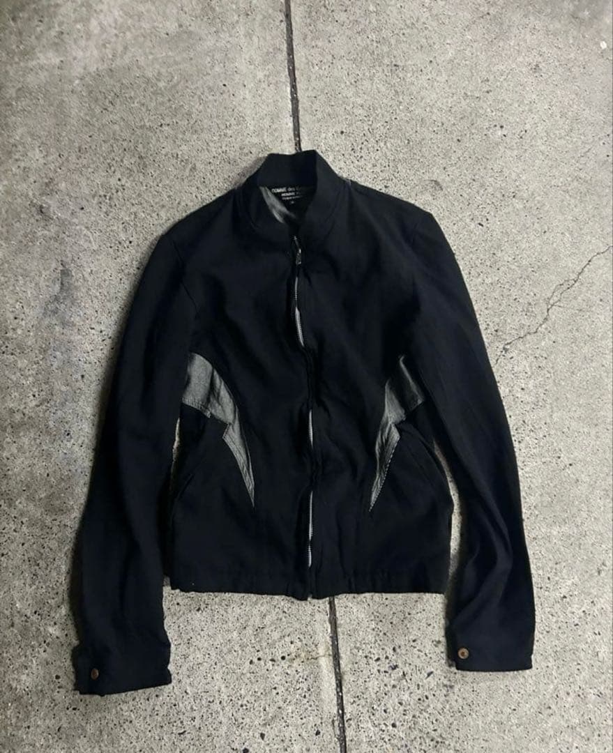 Black COMME des GARCONS Homme Plus men's cotton jacket laid flat on concrete, front view with zipper and lighter side panels visible.
