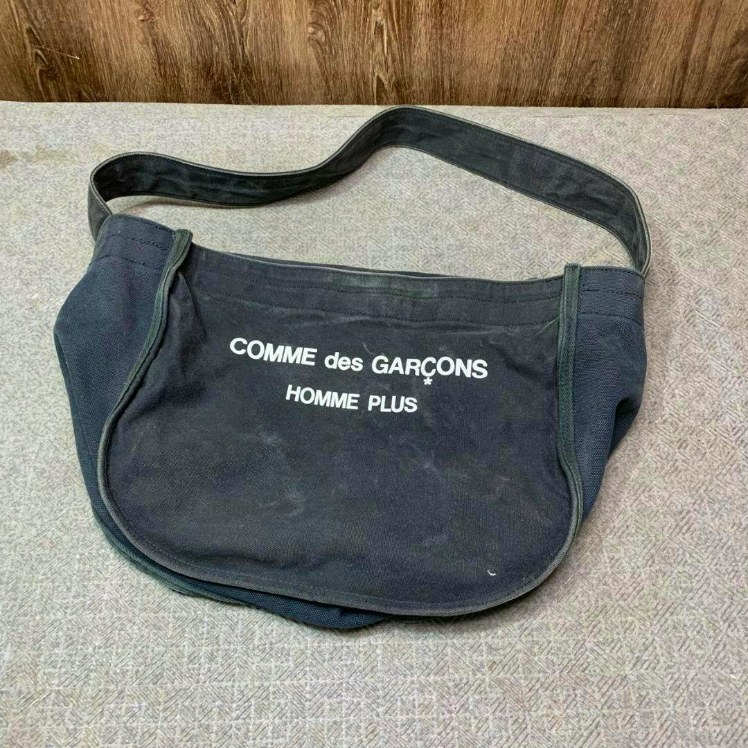 Black canvas shoulder bag with white "COMME des GARÇONS HOMME PLUS" text printed on the front, shown laid flat.