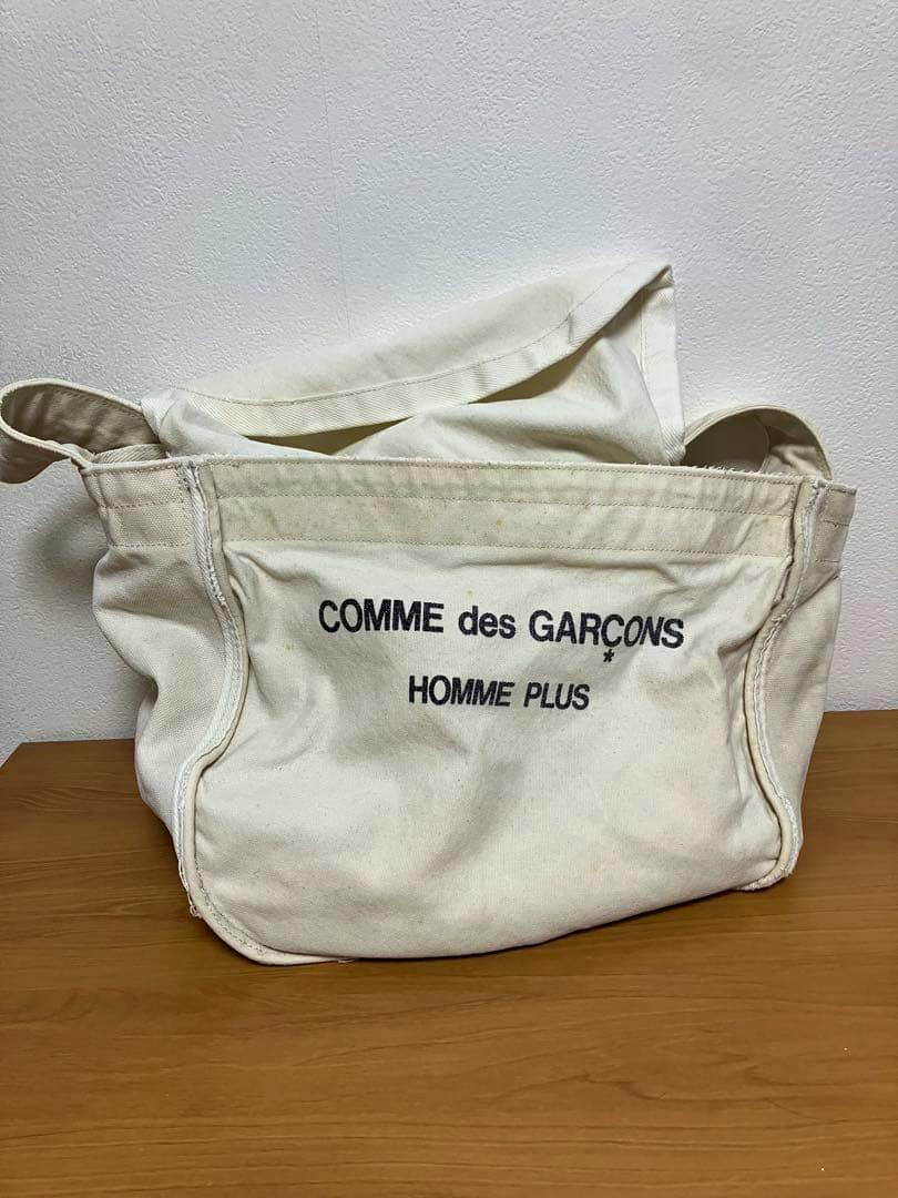 Front view of a white/ivory canvas COMME des GARÇONS HOMME PLUS shoulder bag showing the printed logo.
