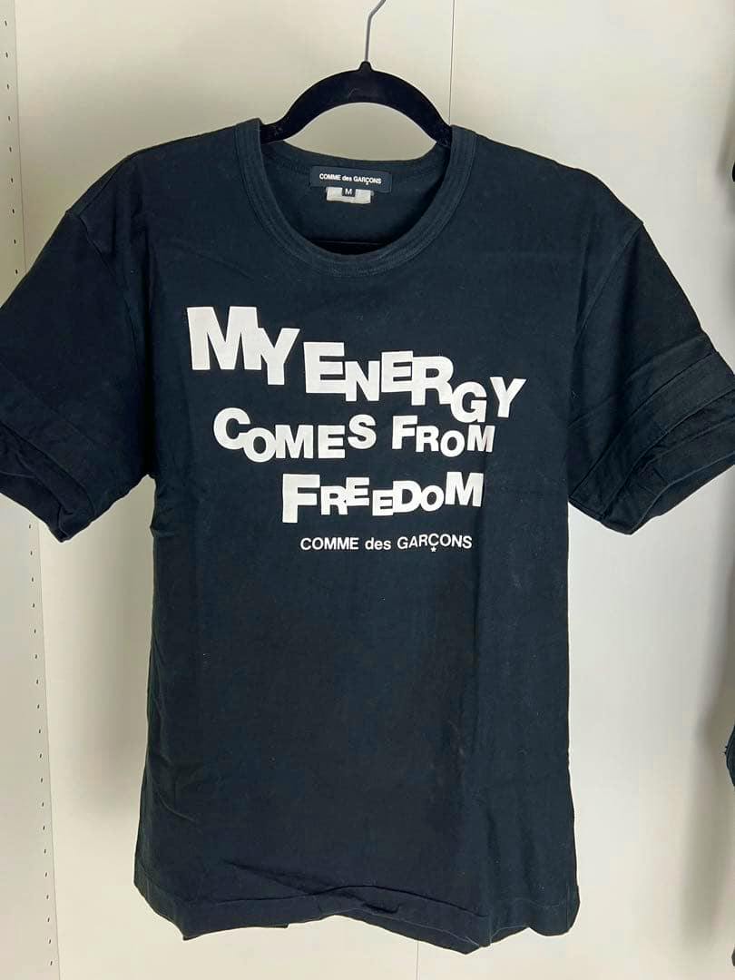 Black COMME des GARÇONS cotton T-shirt (size M) with white printed front text hanging on a hanger.