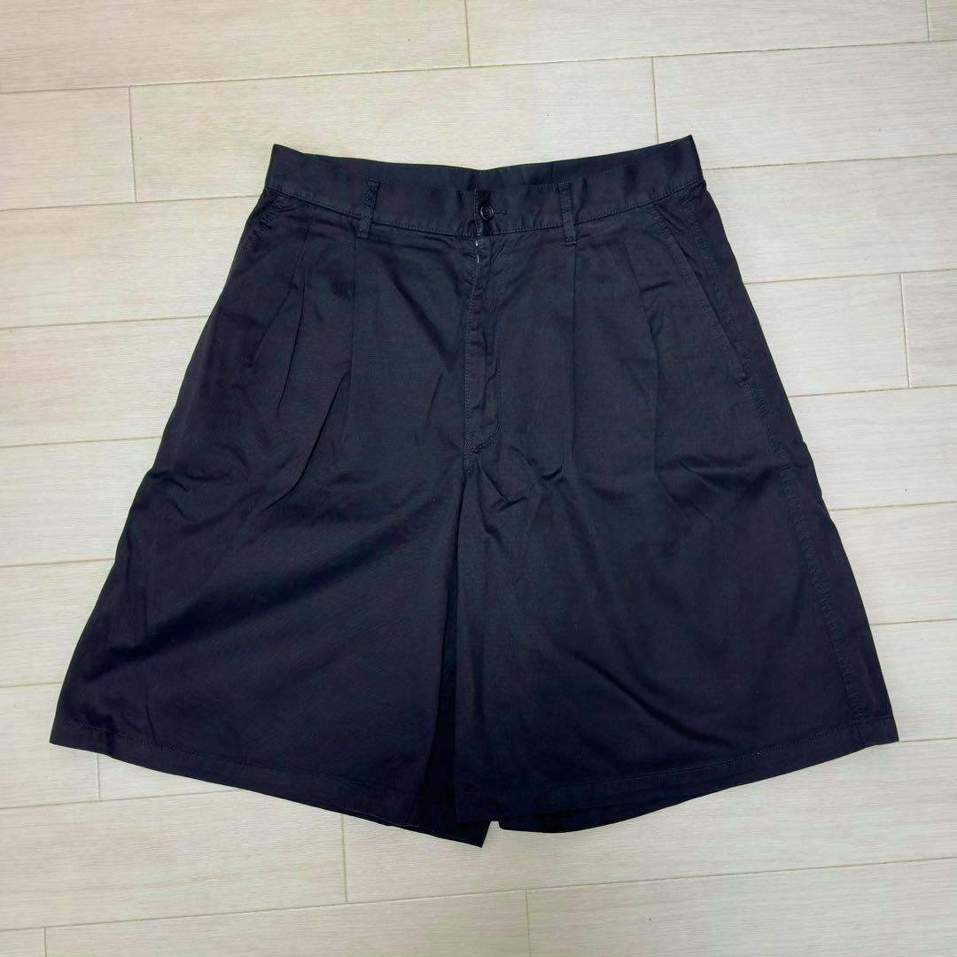 Front view of black COMME des GARCONS SHIRT two-tuck wide men's cotton shorts laid flat on a light wood floor.