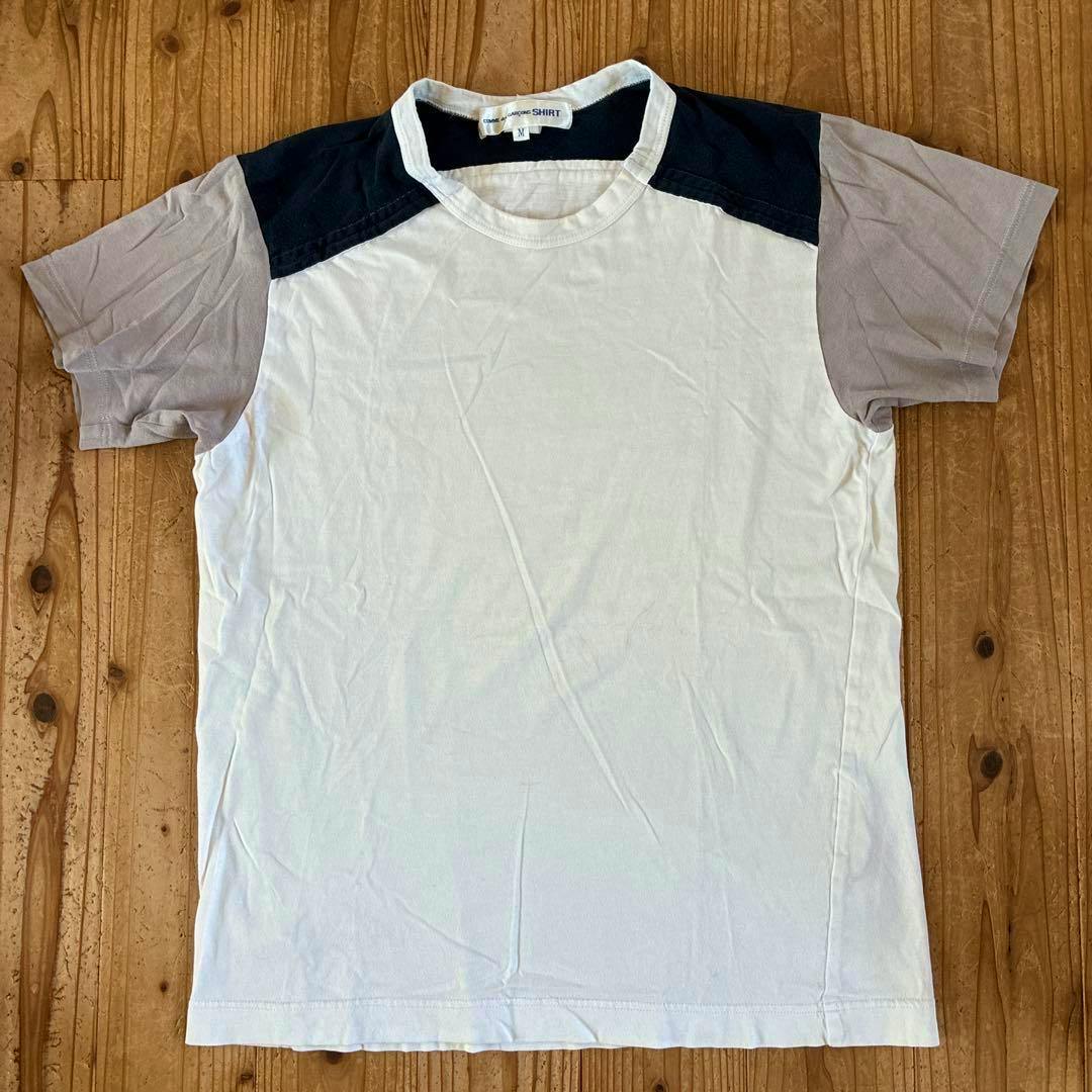 Front view of the COMME des GARCONS SHIRT cotton T-shirt with a dark shoulder yoke and gray sleeves laid flat on a wooden floor.