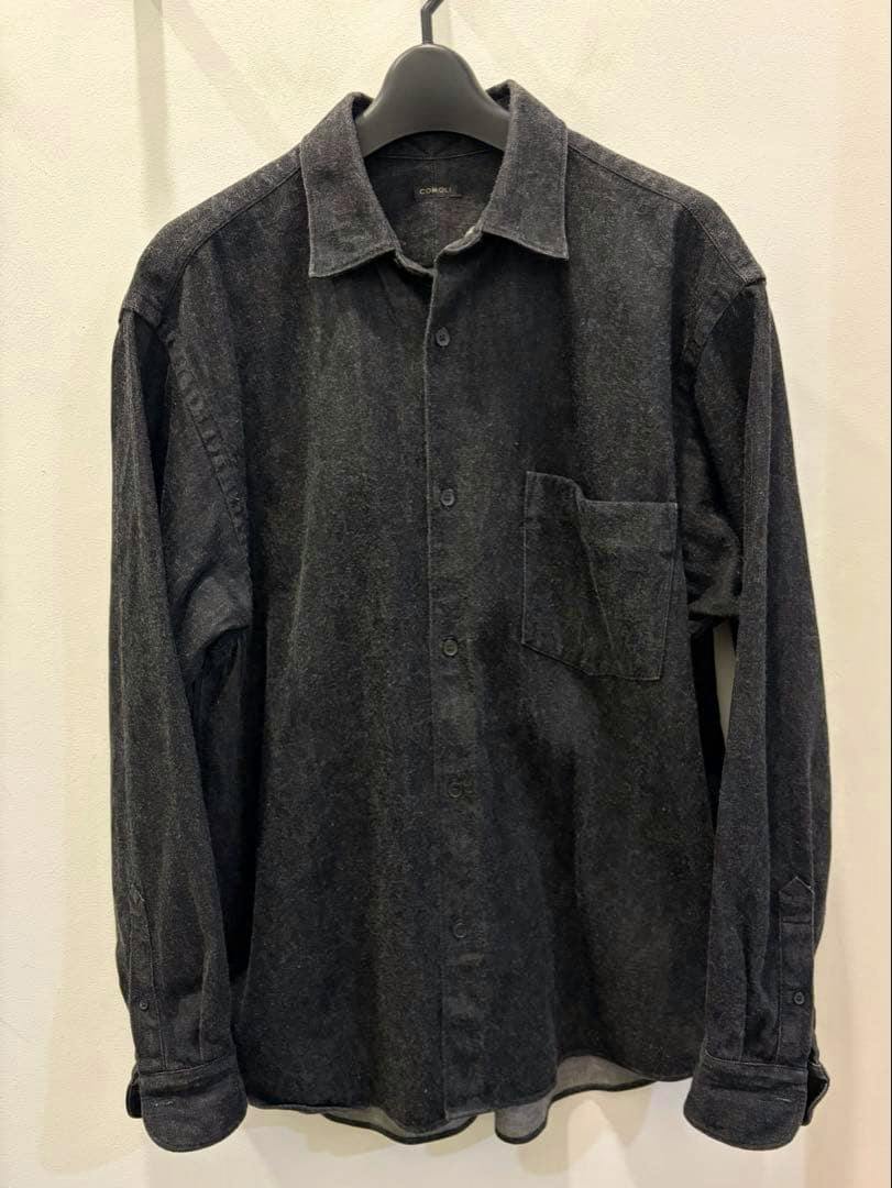 Front view of a dark denim Comoli men's long-sleeve button-up shirt hanging on a hanger against a wall.
