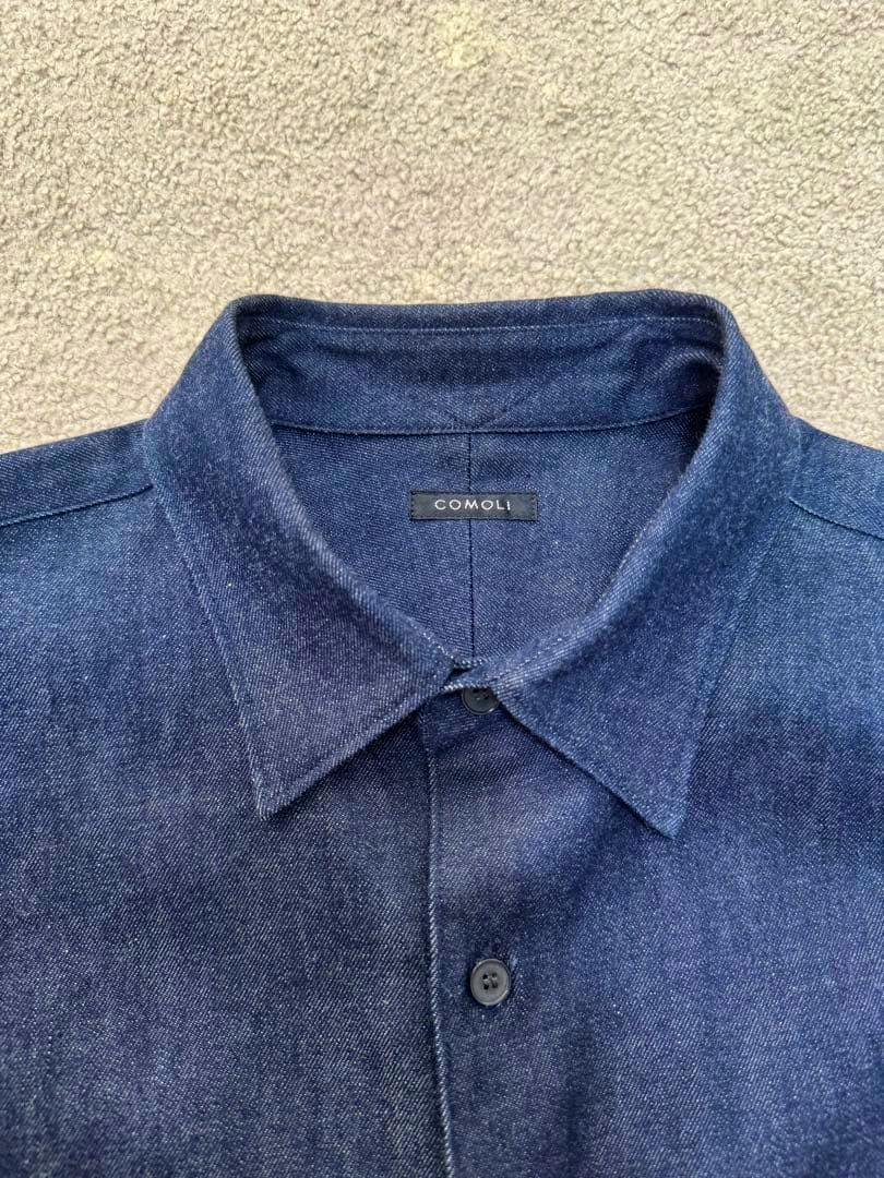 Close-up of the collar, top buttons, and COMOLI label on a blue denim men's shirt.
