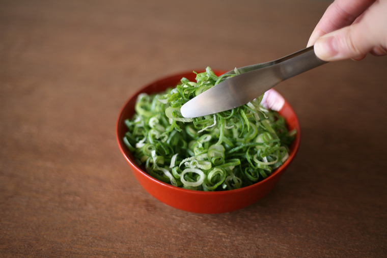 Hand holding metal tongs picking sliced green onions from a red bowl on a wooden surface.