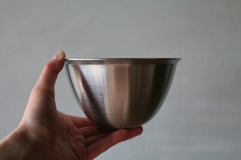 Hand holding an empty metal Conte serving round bat (bowl) against a plain background.