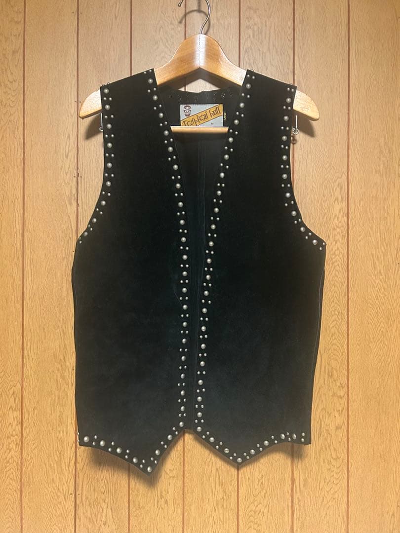 Front view of a black cow leather studded vest (L size) hanging on a wooden hanger against a wood-paneled background.