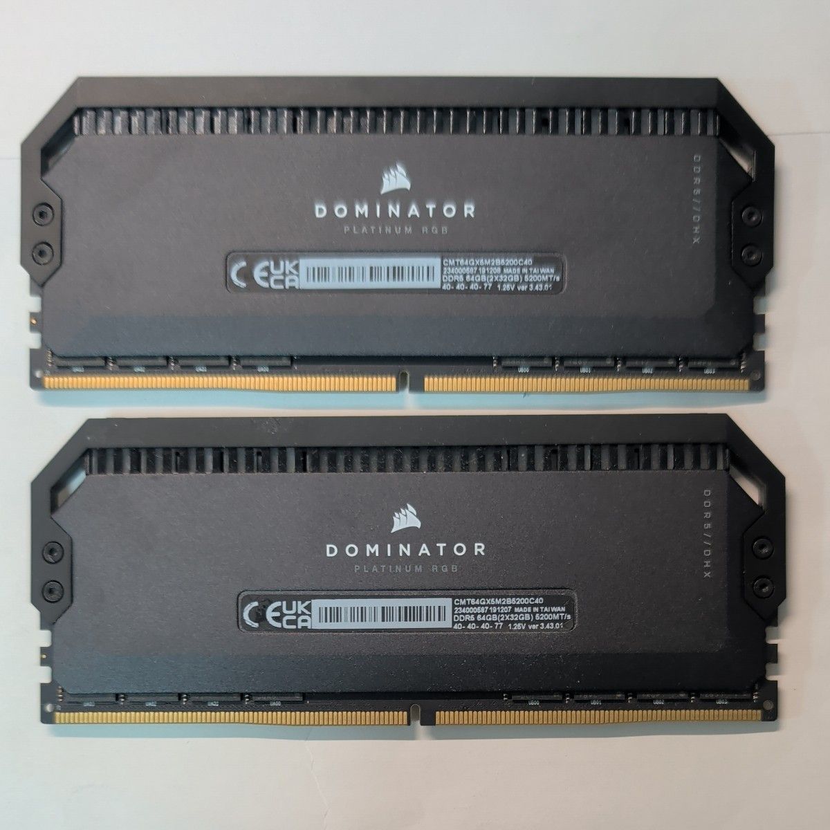 Two CORSAIR DOMINATOR PLATINUM RGB DDR5 RAM modules (32GBx2) laid flat on a light surface, showing their top heatspreaders and gold connector pins.
