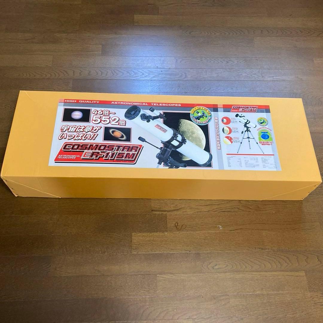 Photograph of the yellow box containing the Cosmostar ER-115M astronomical telescope with its product label visible on a wooden floor.