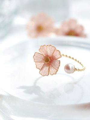 A ring with a pink cosmos flower and a small pearl on a twisted band photographed on a white surface.