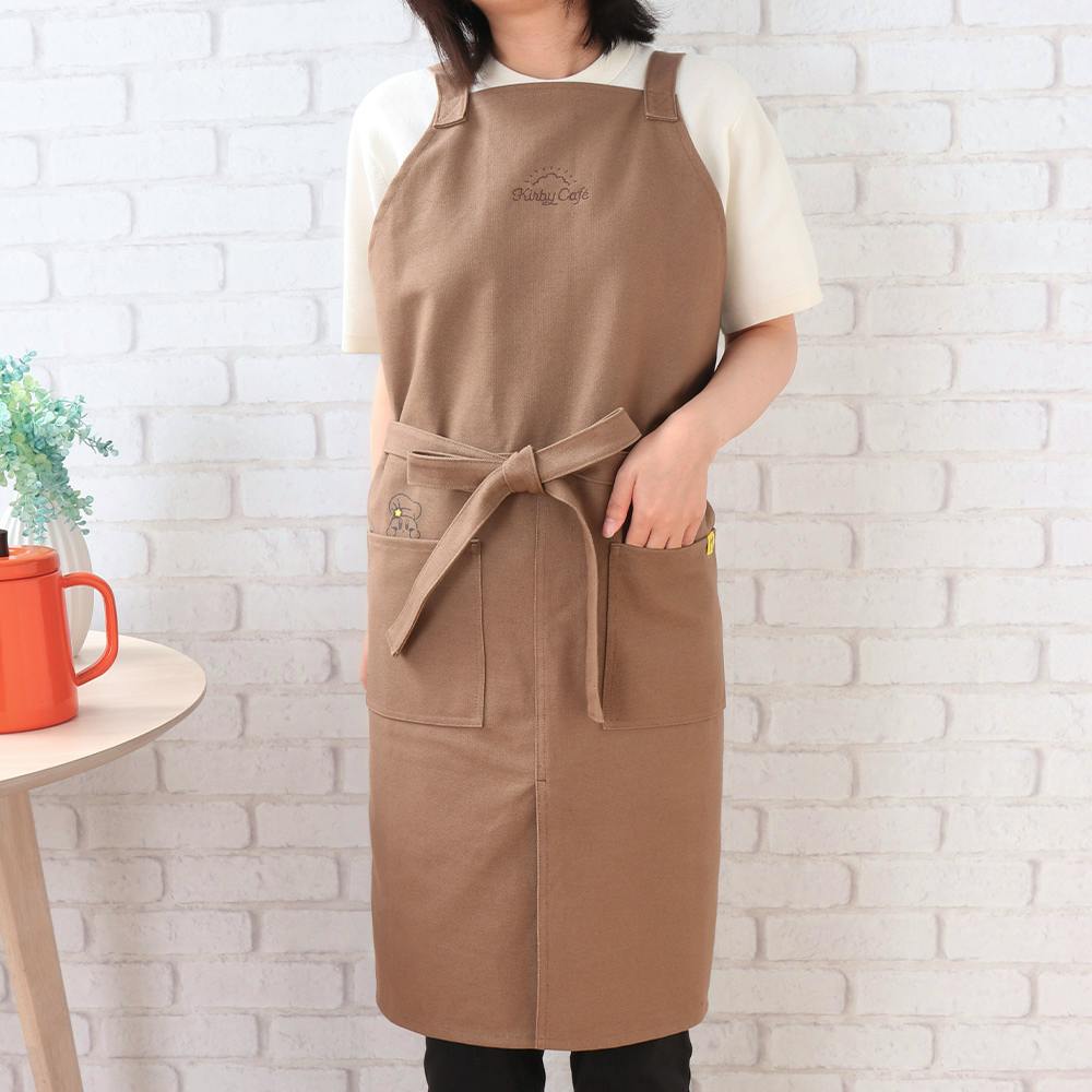 A brown cotton apron with front pockets and a tied waist being worn by a person standing against a white brick wall.