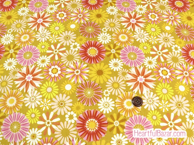 Flat spread of cotton fabric showing a yellow background with multicolored daisy-like floral pattern and a brown button for scale.