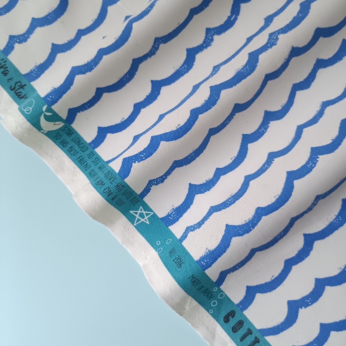 A piece of Kujira and Star cotton fabric showing a repeated blue wave pattern and a teal selvedge with text.