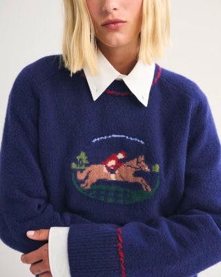 Model wearing a navy cotton sweater with a horse-and-rider embroidered motif on the front.