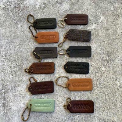 Multiple rectangular cow leather hang tag keychains with ball chains in various colors arranged on a textured surface.
