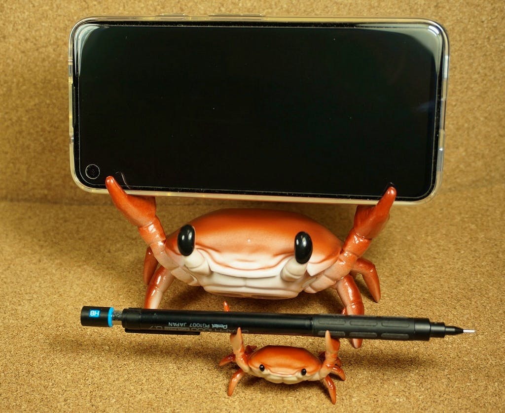 A crab-shaped phone holder supports a smartphone while a smaller crab-shaped pen holder holds a pen in front.