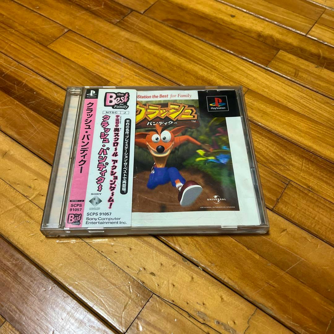 Front cover of the Japanese "Best" edition Crash Bandicoot PlayStation game in a jewel case with an obi strip, shown on a wooden floor.