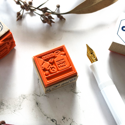 Square orange rubber stamp mounted on a wooden block shown with a fountain pen nib and a dried sprig on a marble surface.