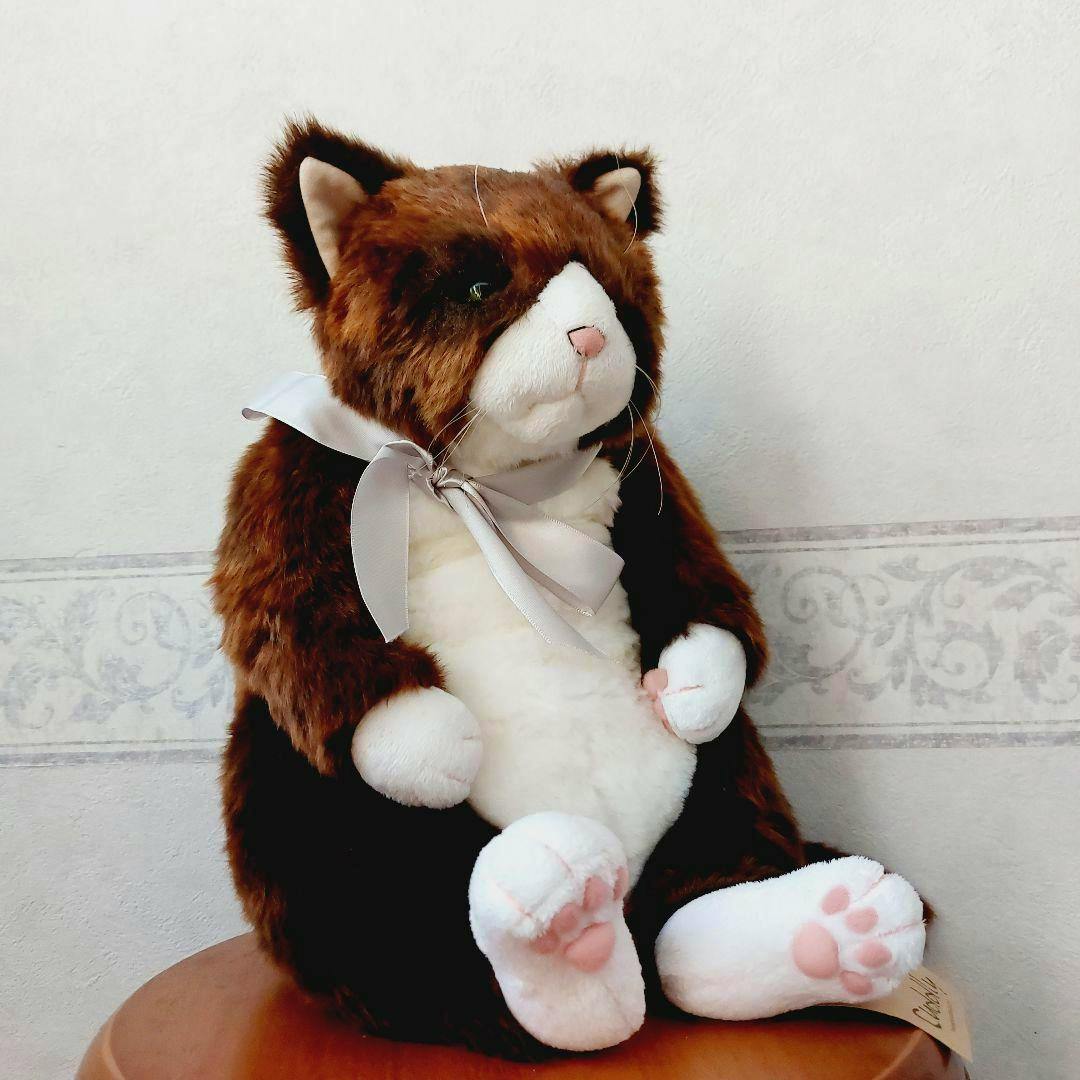 A brown-and-white plush toy cat wearing a gray ribbon bow and sitting on a surface.
