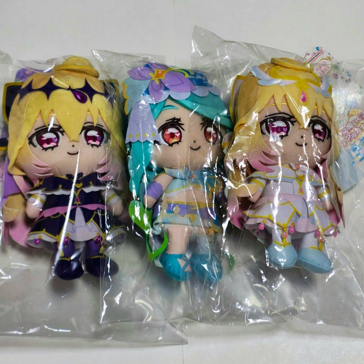 Three plush dolls from the listed 3-piece set (キュアアルカナシャドウ, キュアエクレール, キュアアルカナ) shown in clear plastic packaging on a white surface.