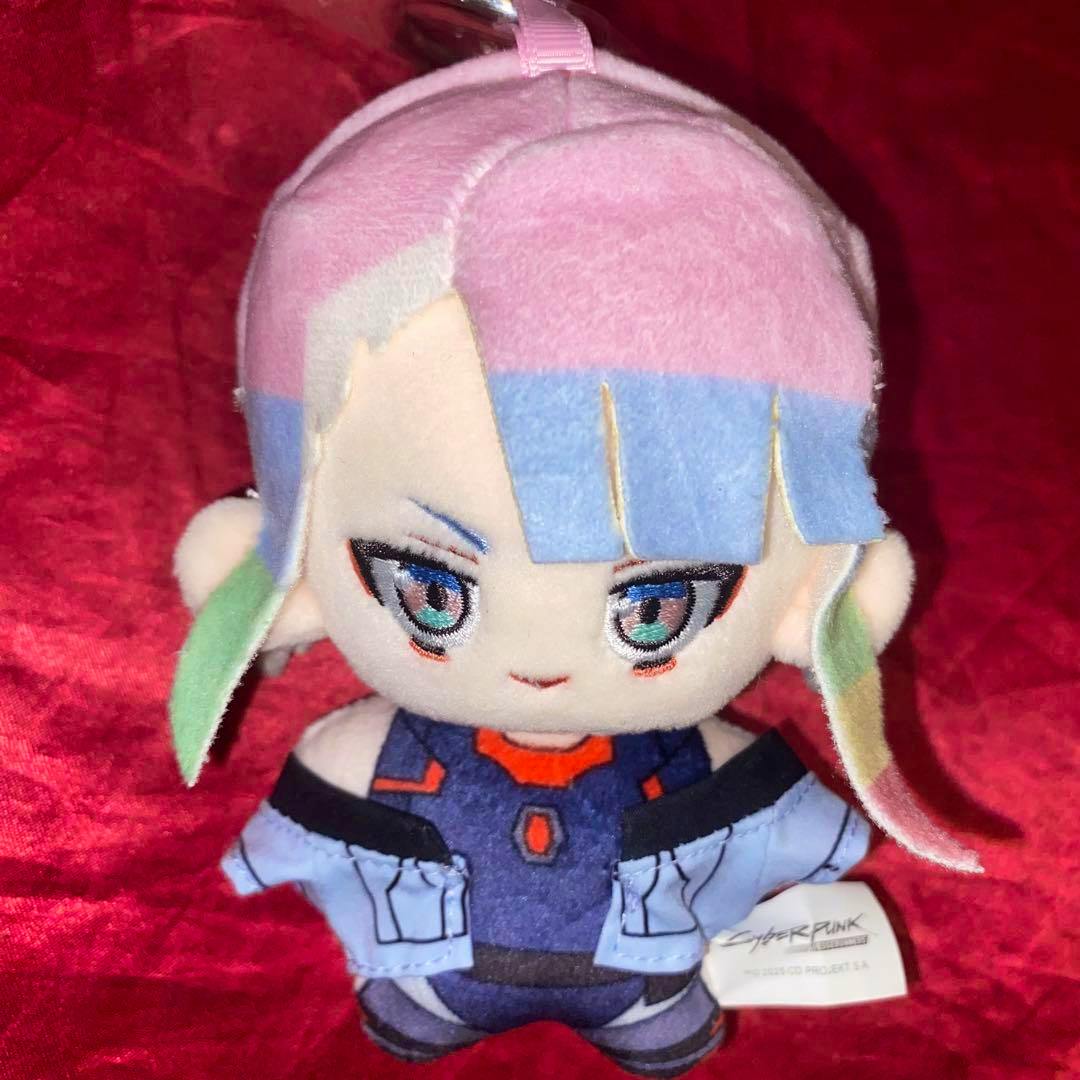 Front view of the Cyberpunk Edgerunners Lucy plush toy showing pink and blue hair, embroidered eyes, and her outfit on a red background.