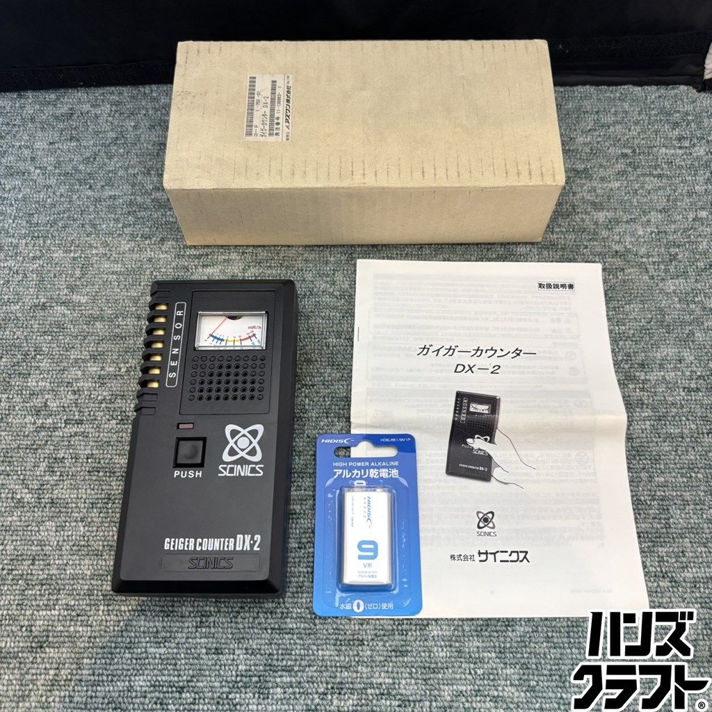 DX-2 Geiger counter (radiation detector) shown with its instruction manual, a packaged battery, and the cardboard box on a carpeted surface.