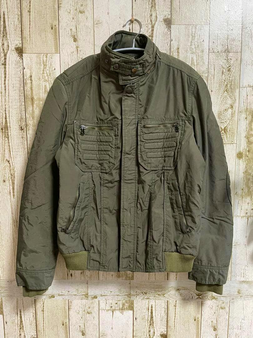 Olive green D&G military jacket hanging on a hanger against a wooden plank background.