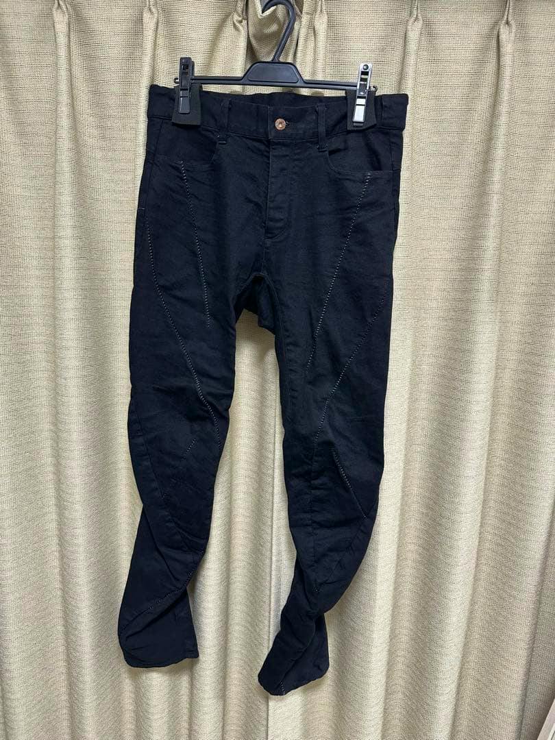 Front view of black men's pants hanging on a hanger against a beige curtain.