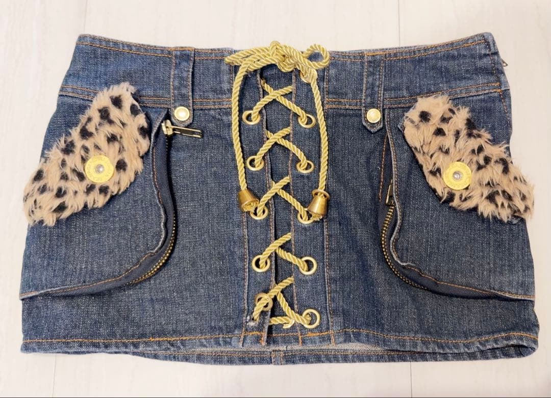 Flat-lay front view of a denim mini skirt with a center lace-up detail and leopard-print fur pocket accents.