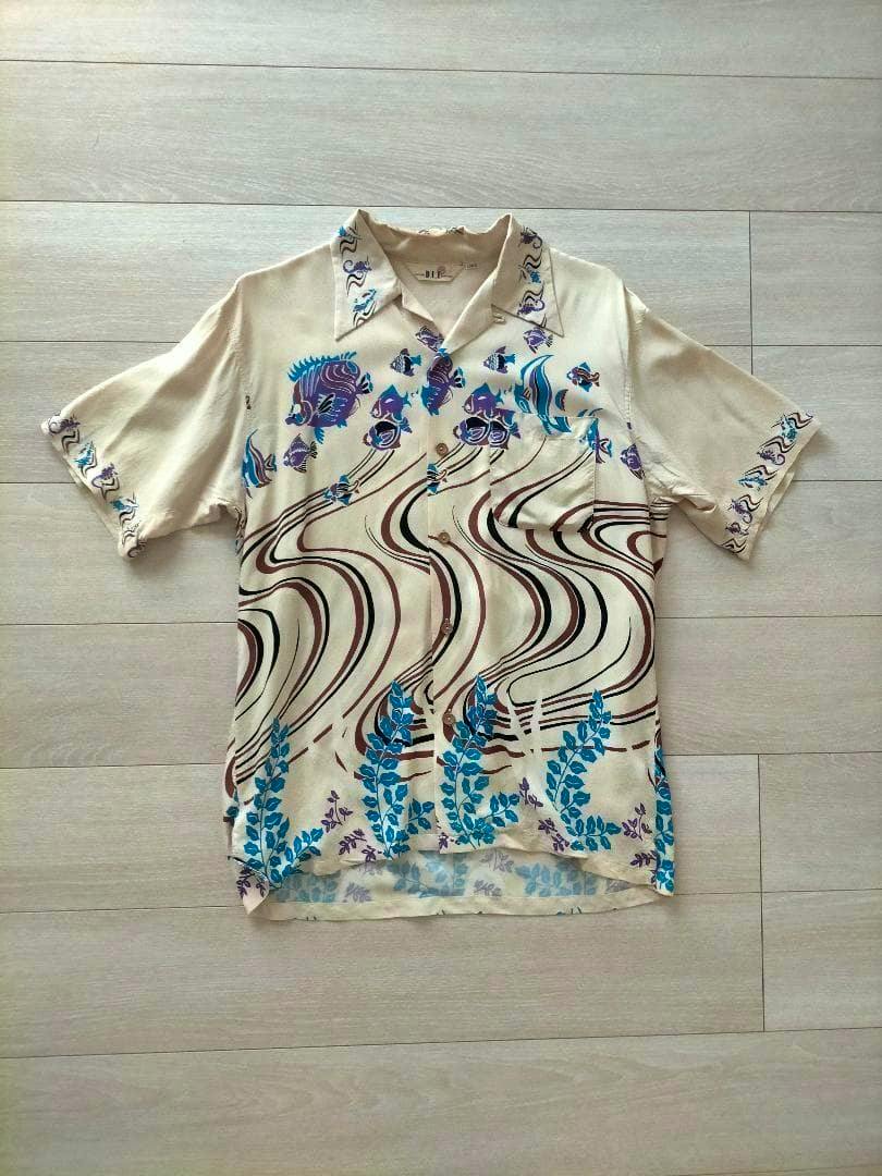 Beige short-sleeve vintage Aloha shirt with blue leaf and purple fish motifs and brown wavy lines laid flat on a light wood floor.