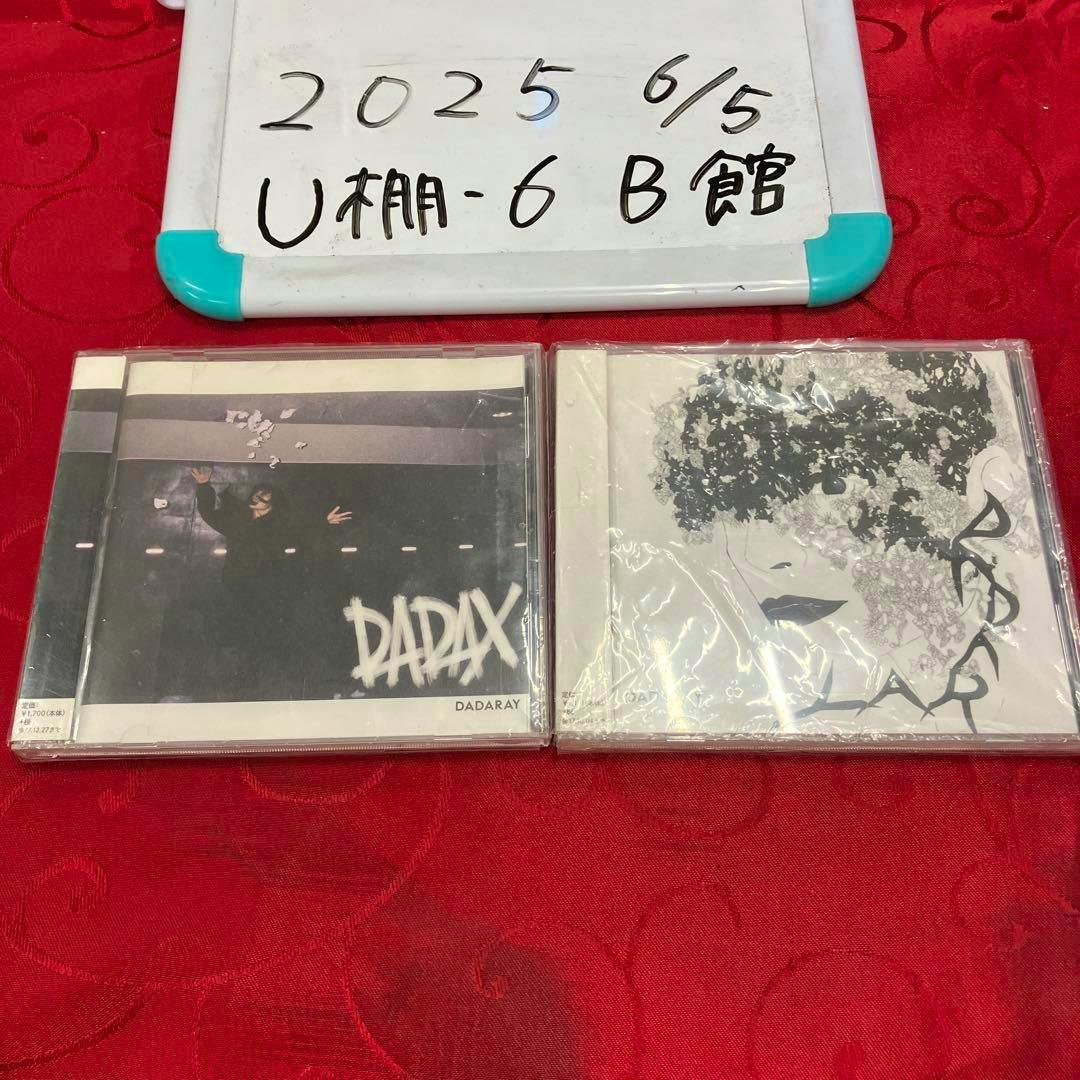 Two DADARAY CD cases shown side-by-side on a red cloth surface with a whiteboard behind them.