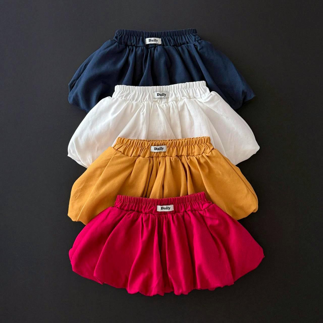Four children's pumpkin-style skirts in navy, white, mustard, and bright pink stacked on a dark background, each showing a small 'Daily' label.