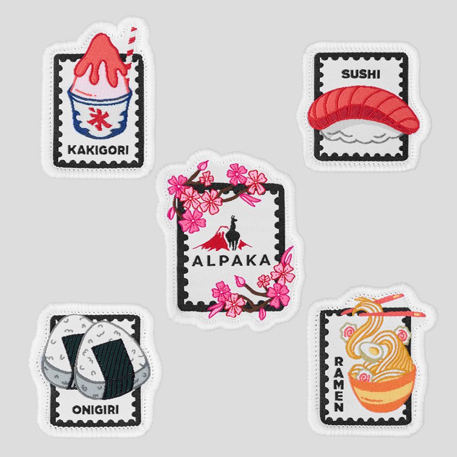 Five embroidered fabric patches laid out on a gray background showing kakigori, sushi, onigiri, ramen, and a central ALPAKA cherry-blossom design.