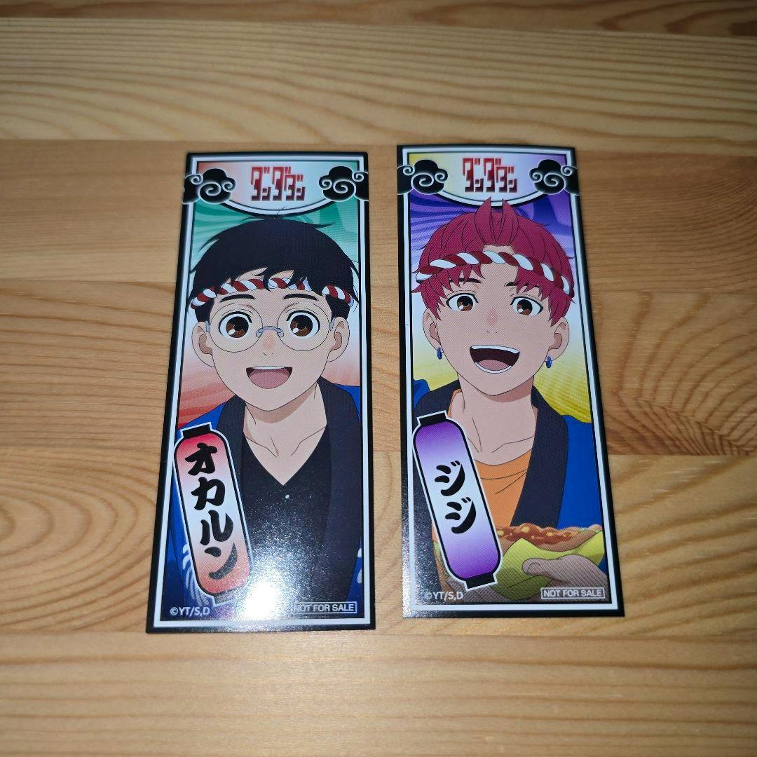 Photo of two rectangular stickers on a wooden surface featuring the characters オカルン and ジジ from ダンダダン.