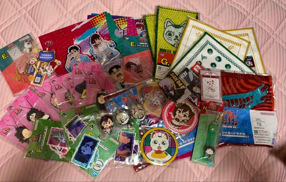 A collection of Dandadan Ichiban Kuji goods including acrylic stands, can badges, and towels spread on a pink quilted surface.