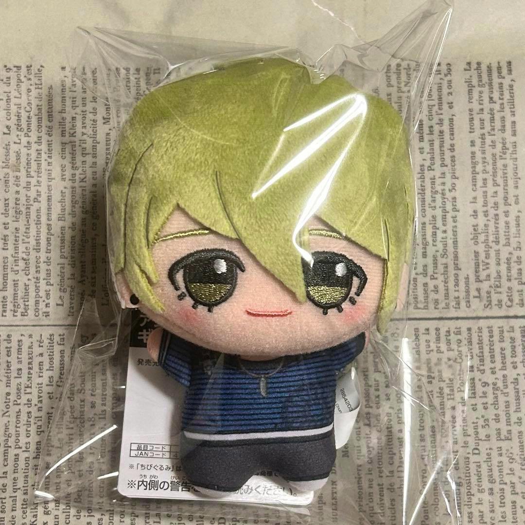 A plastic-wrapped chibi plush of Rantaro Amami from Danganronpa, showing light greenish-blond hair, embroidered eyes, and a blue striped outfit on a newspaper background.